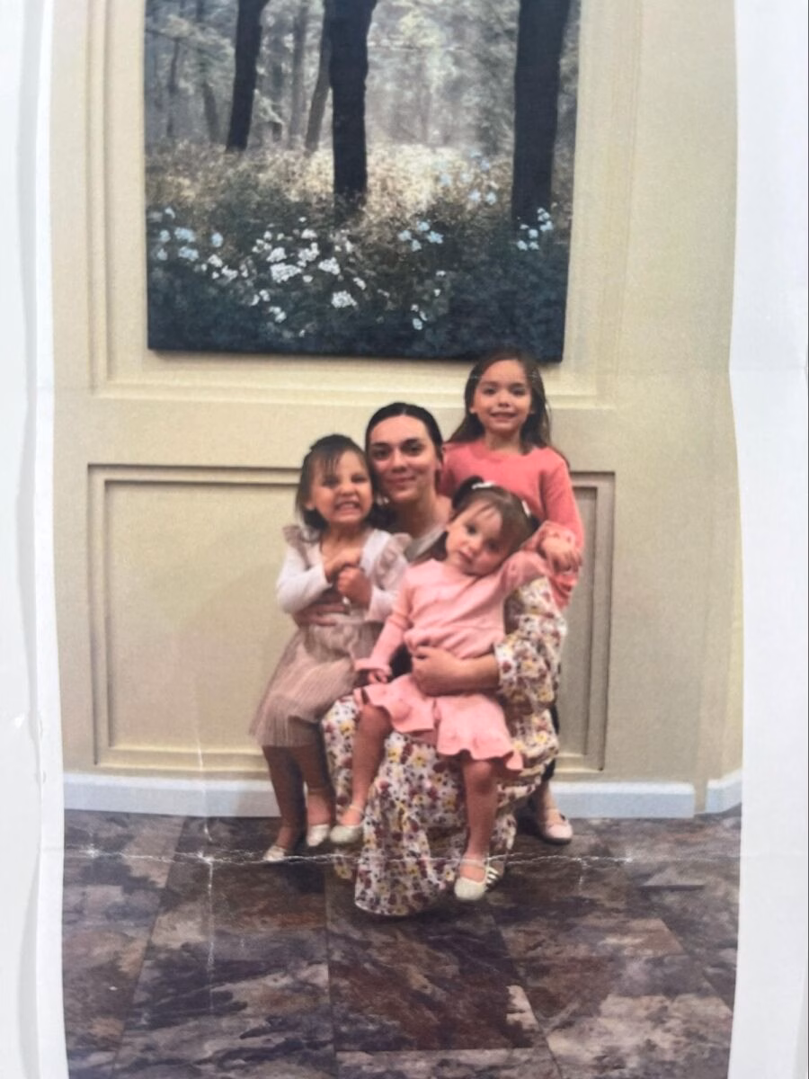 A woman in a floral dress sits on a chair with three young girls. One girl is on her lap, another sits beside her, and a third stands behind her, all smiling in front of a forest painting.