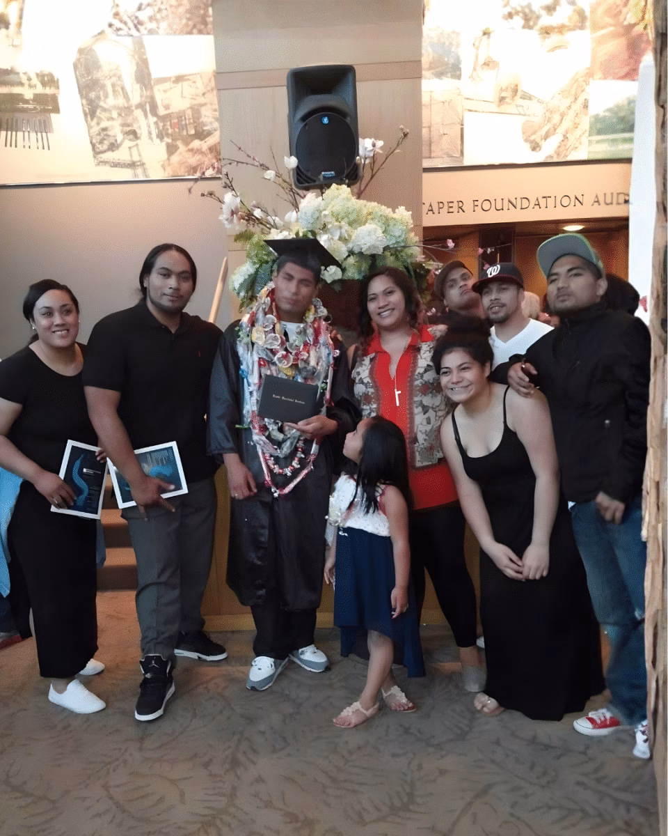 A group of nine people, including a graduate in a cap and gown with a lei, posing for a photo in front of a sign that reads 'TAPER FOUNDATION AUD'.