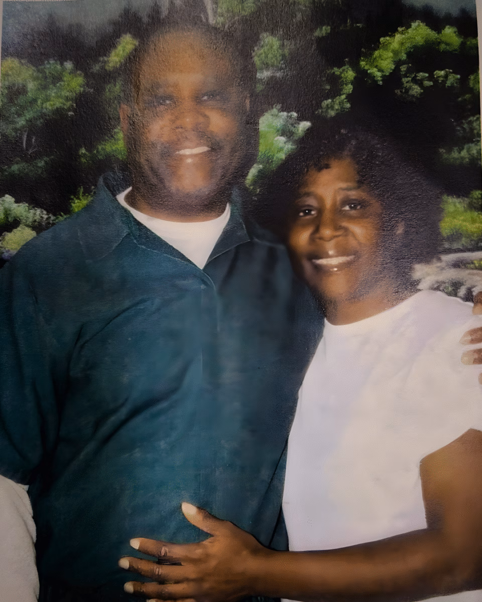 3 A portrait of a smiling couple, Charles and his partner, standing together in front of a lush green forest backdrop.