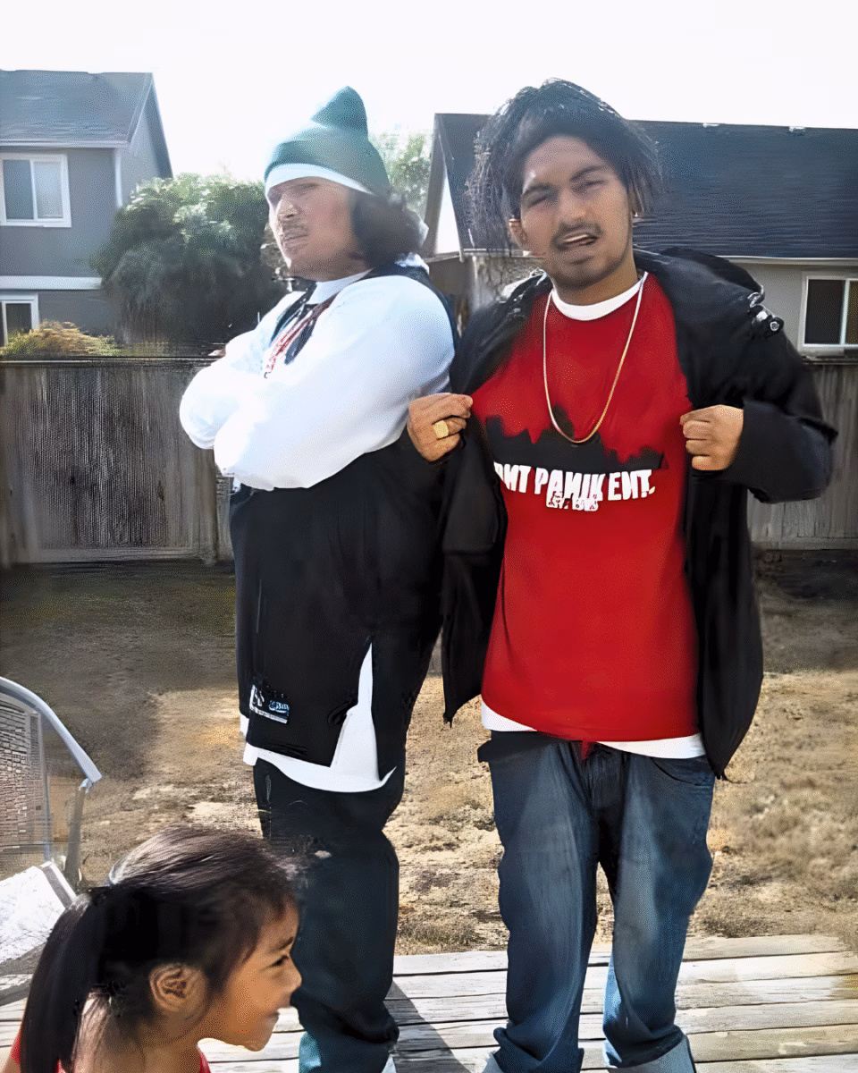 Two men stand on a wooden deck outdoors; one wears a green beanie and white shirt, the other a red shirt with 'PAMIK EXT.' text, with a young girl in the foreground.