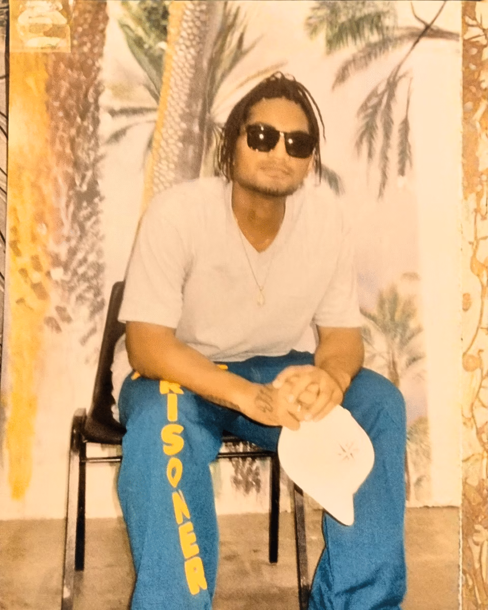 A man with dreadlocks and sunglasses sits on a chair, wearing blue pants with 'RISONEER' in yellow and holding a white cap, against a palm tree mural.