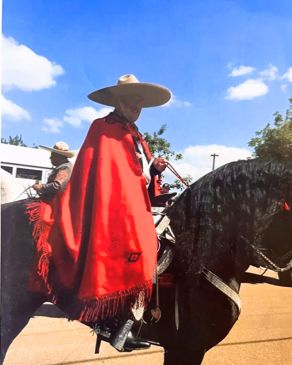 A man in a red poncho and wide-brimmed hat rides a black horse under a blue sky.