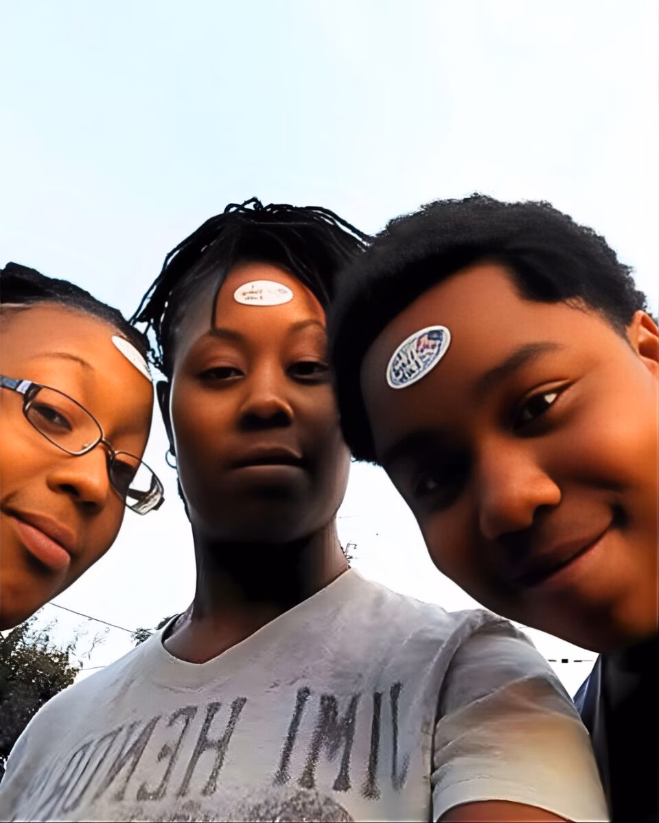 Three young people posing for a selfie, each wearing a sticker on their forehead, against a bright sky.