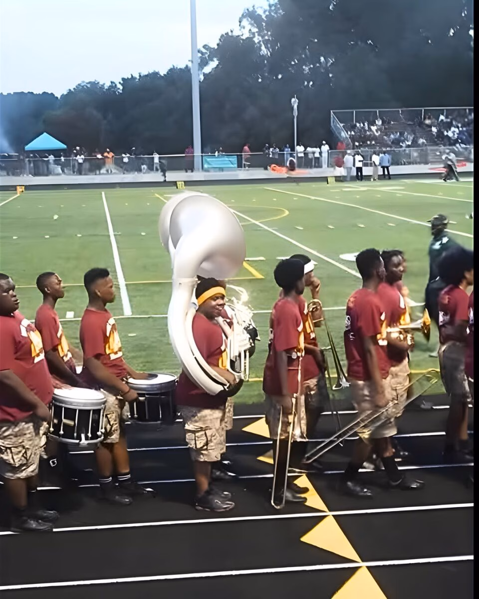 A marching band in maroon shirts and camouflage shorts performs on a football field, featuring a large tuba player in the center.