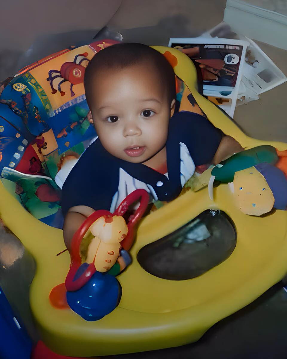 Diyondré Wilson, a young child, looks forward from a yellow activity center with colorful toys.