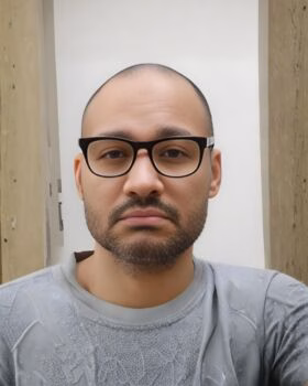 Portrait of Raymond, a man with a shaved head, wearing black-rimmed glasses and a grey t-shirt.