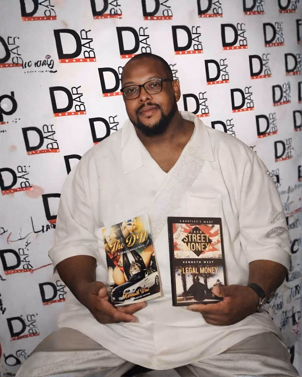 Kenneth West (1) Kenneth West holds two of his books, 'The DM' and 'From Street Money to Legal Money', in front of a D-BAR event backdrop.