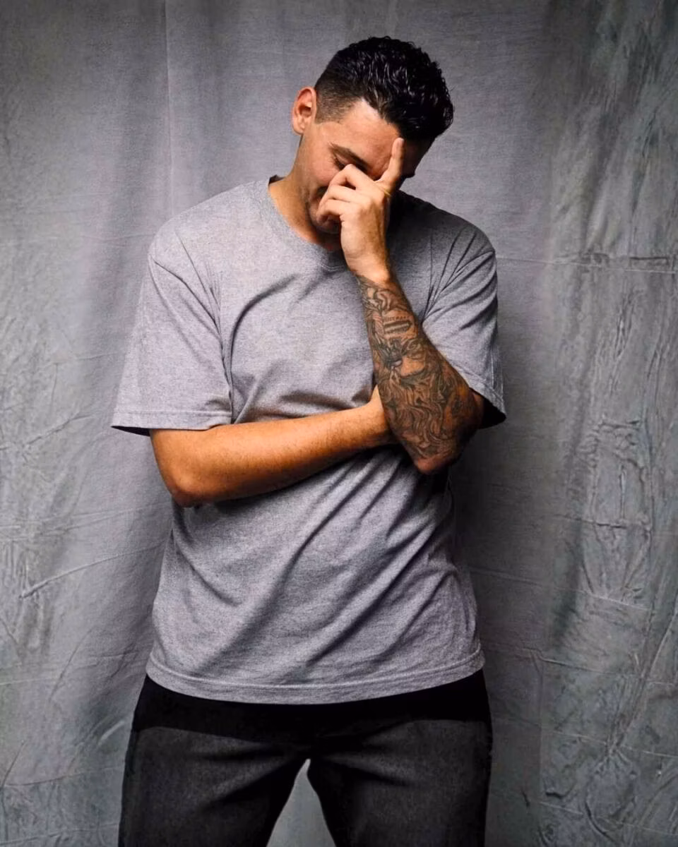 Robert Ortiz (3) Robert Ortiz, 32, wearing a grey t-shirt and dark pants, stands against a grey backdrop with his hand covering his face. He has a detailed tattoo on his left forearm.