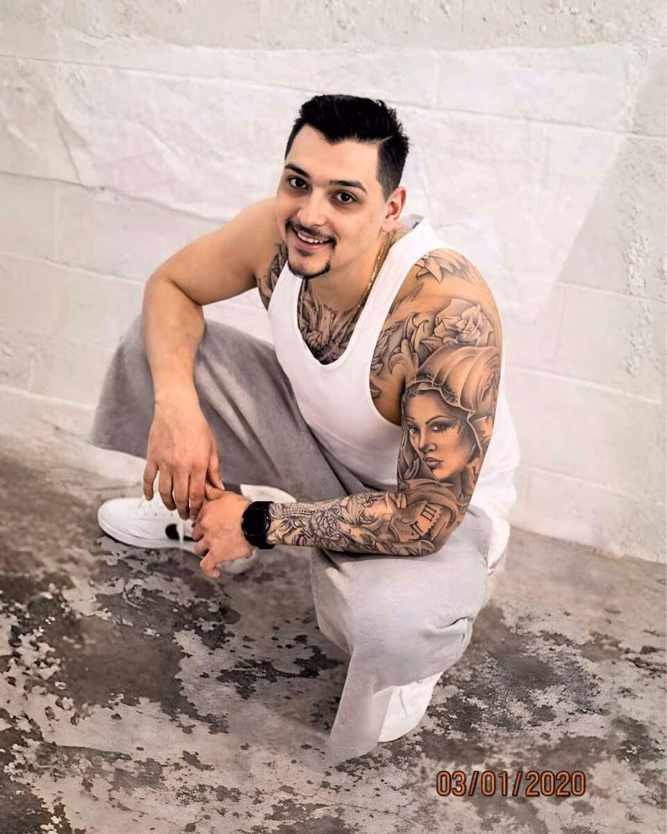 Robert Ortiz (4) Robert Ortiz crouching against a white brick wall, wearing a white tank top and grey sweatpants, with extensive tattoos on his arms.