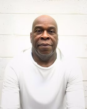 Portrait of Allen Lynn Penn, a bald Black man with a white beard, wearing a white long-sleeved shirt against a white brick wall.