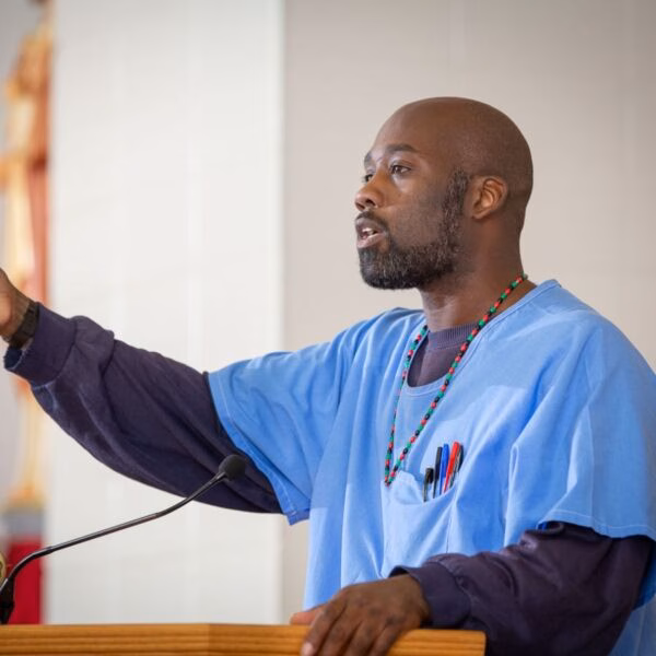 George 'Mesro' Coles, a man in blue scrubs, speaks at a podium with a microphone.