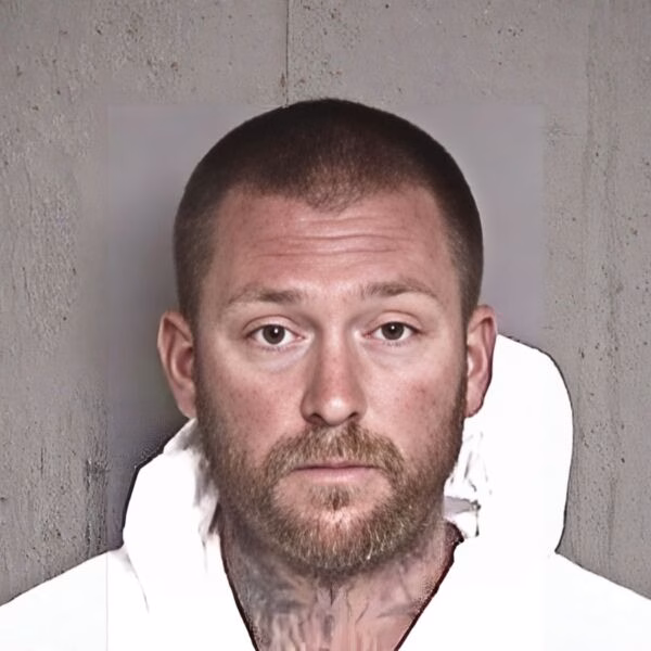 Mugshot of Greg Stephens, a man with a short haircut and beard, wearing a white shirt against a concrete wall.