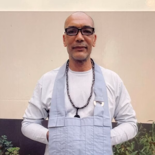 Mark Cádiz, a bald man with glasses, wearing a light blue apron over a white shirt and a beaded necklace, stands against a plain wall.