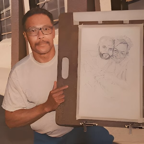 Mark Stanley, an artist with glasses and a mustache, points to a pencil sketch of himself and another person on an easel.