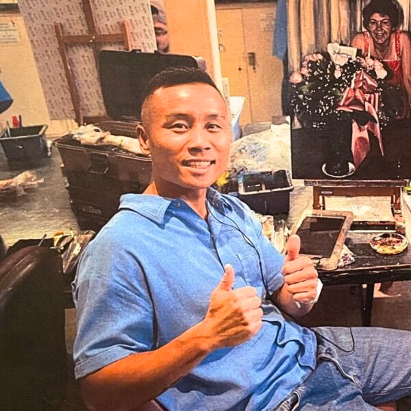 Peter Thao, an artist, gives two thumbs up in his studio, with a portrait of a woman on an easel behind him.