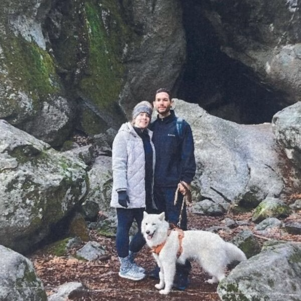 Justin COOKSEY and a companion stand with a white dog in front of a mossy rock cave.