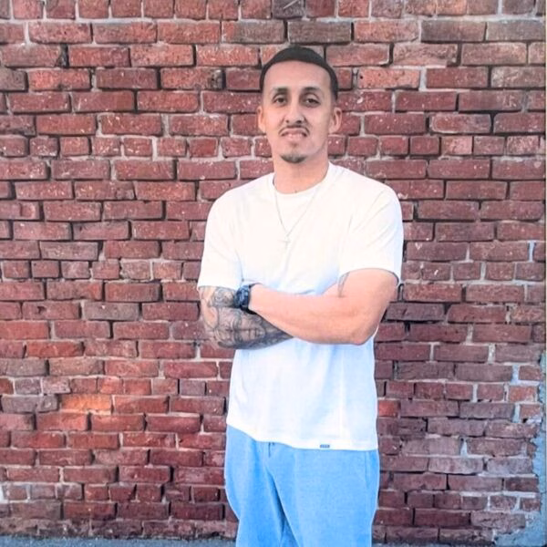Michael Navarro stands with arms crossed in front of a red brick wall, wearing a white t-shirt and light blue pants.