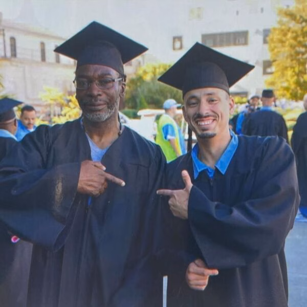 Tony de Trinidad and a young man in graduation gowns both pointing at each other, smiling outdoors.