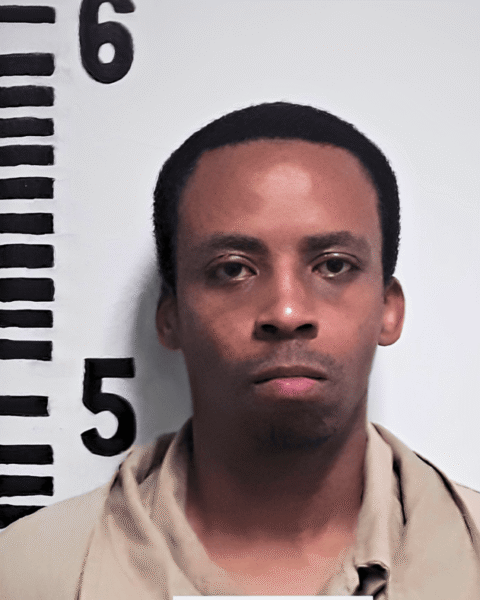 Front-facing portrait of an adult man standing against a height measurement backdrop with black ruler markings and numbers. He has short dark hair, a neutral facial expression, and is wearing a light beige collared shirt. The image resembles an identification or booking photograph