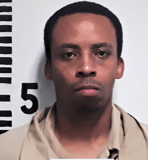 Front-facing portrait of an adult man standing against a height measurement backdrop with black ruler markings and numbers. He has short dark hair, a neutral facial expression, and is wearing a light beige collared shirt. The image resembles an identification or booking photograph