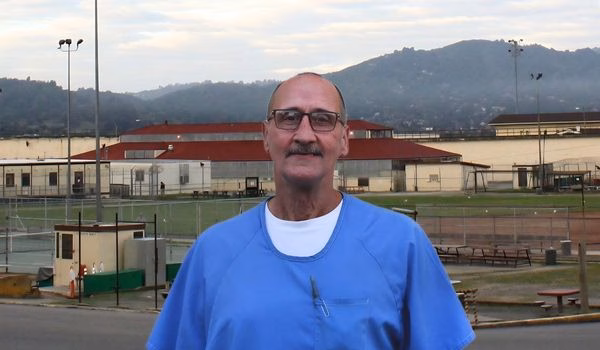 MichealT, a man with glasses and a mustache in blue scrubs, stands in front of a sports facility with hills in the background.