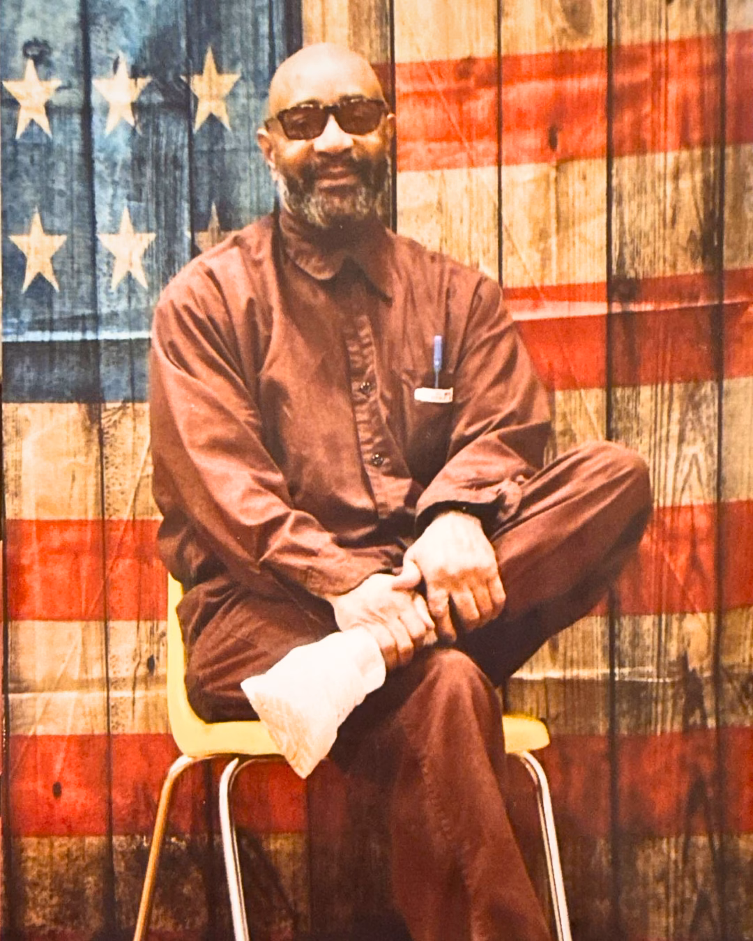 Jeffery, 63, a man with a beard and sunglasses, sits on a chair with his left arm in a cast, in front of a wooden wall decorated with an American flag.