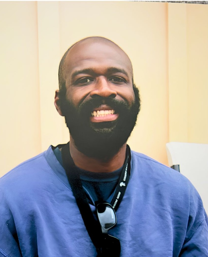 A smiling man with a beard wearing blue scrubs and a lanyard with sunglasses hanging from it.