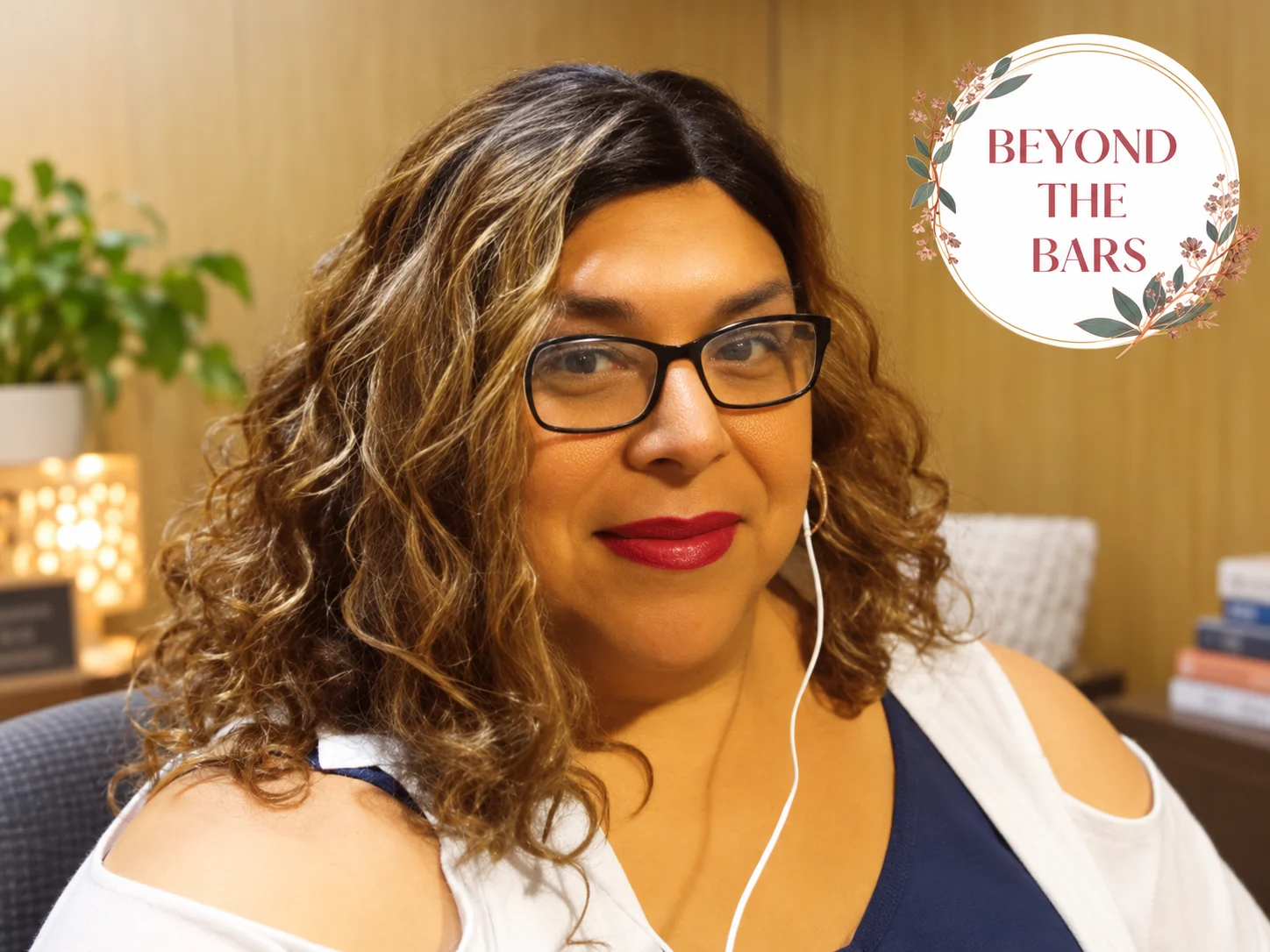 Portrait of Adriel, 26, wearing black-rimmed glasses, red lipstick, and white earphones, with wavy brown hair.
