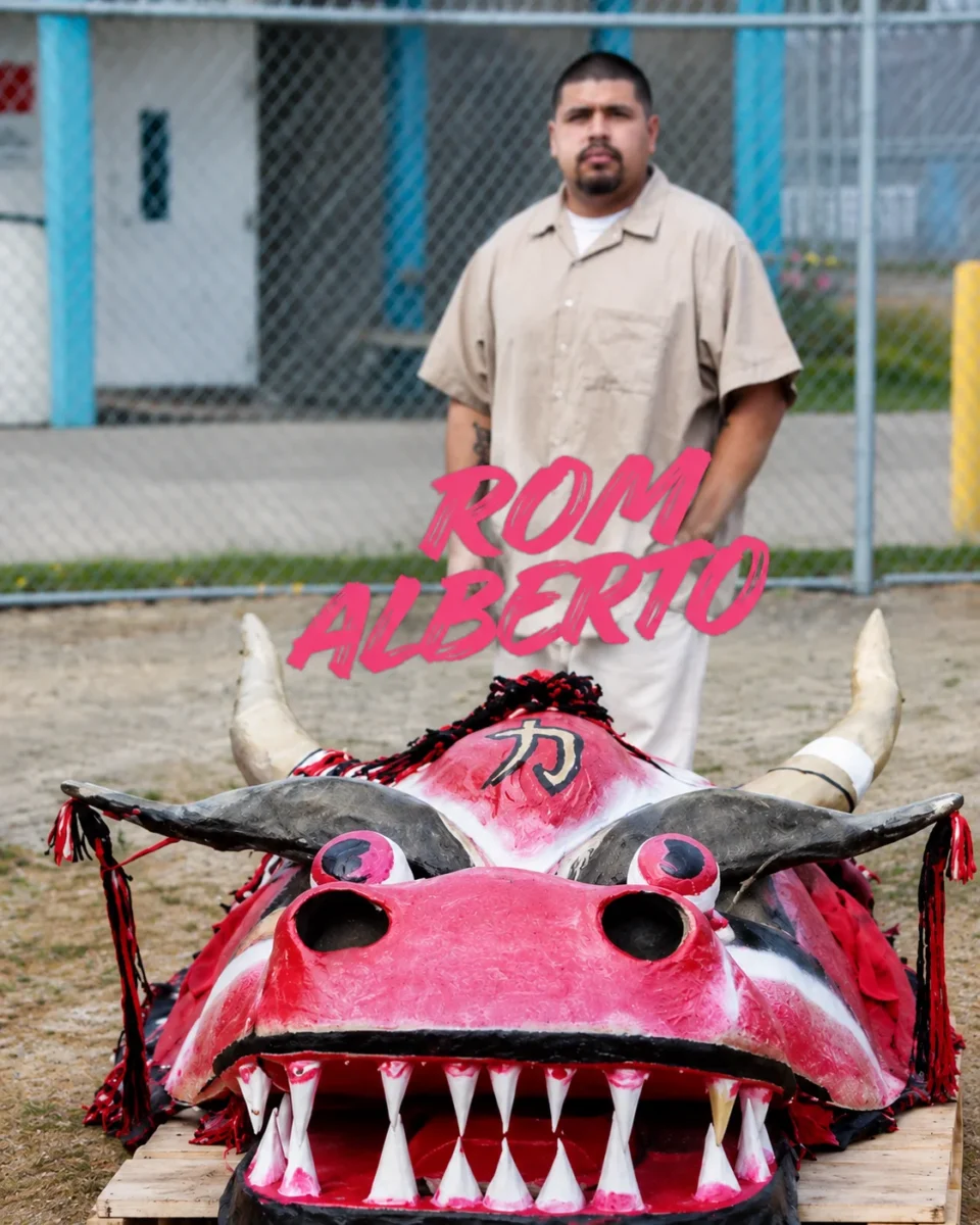 Rom Alberto standing behind a colorful horned artwork in an outdoor prison yard.