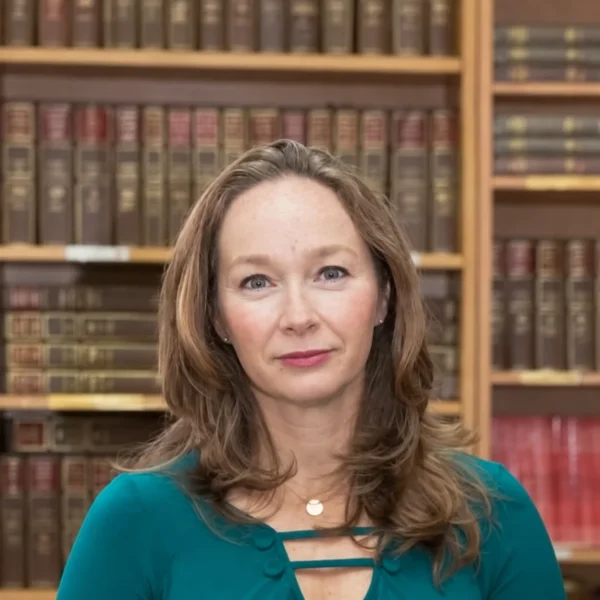 Our Team 16 Andrea, a woman with long brown hair wearing a teal top, stands in front of a bookshelf filled with books.
