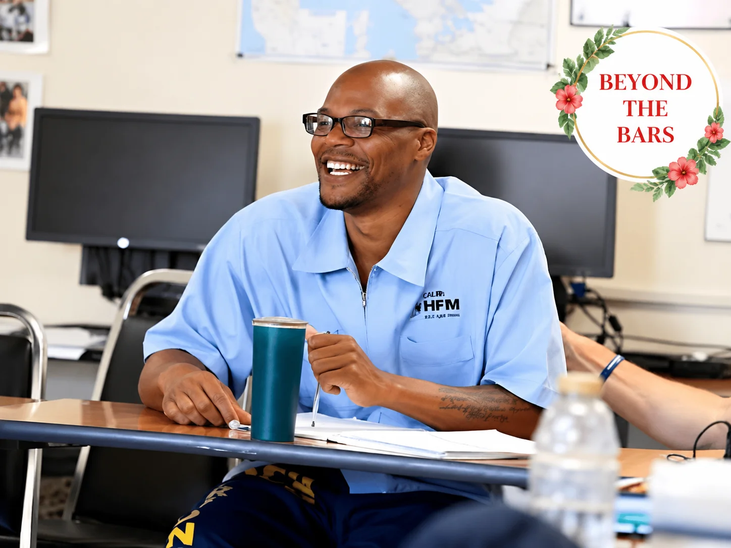 Anthony "Ant" Ammons, wearing a light blue HFM uniform and glasses, smiles while sitting at a desk with a green cup.