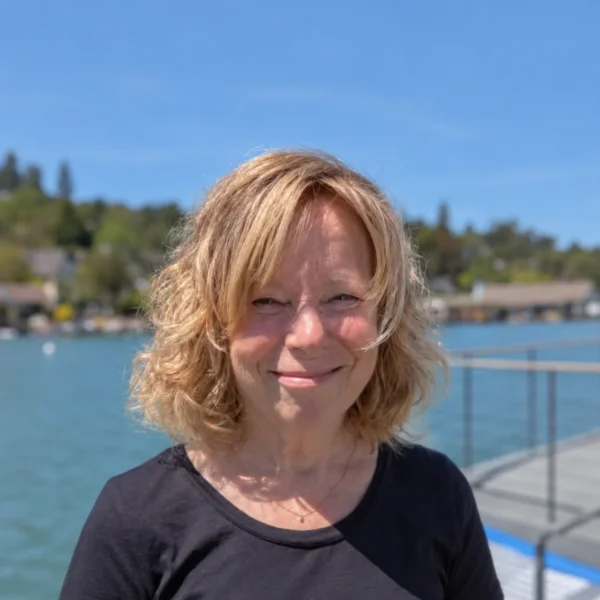 Our Team 15 Portrait of Susan, a woman with blonde hair, smiling outdoors by a body of water.