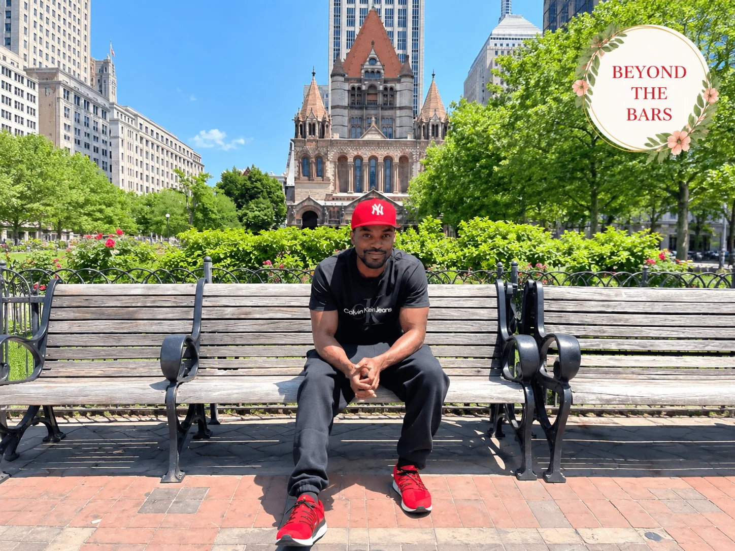 David Baxter sits on a park bench in front of Trinity Church, wearing a red NY cap and Calvin Klein shirt.