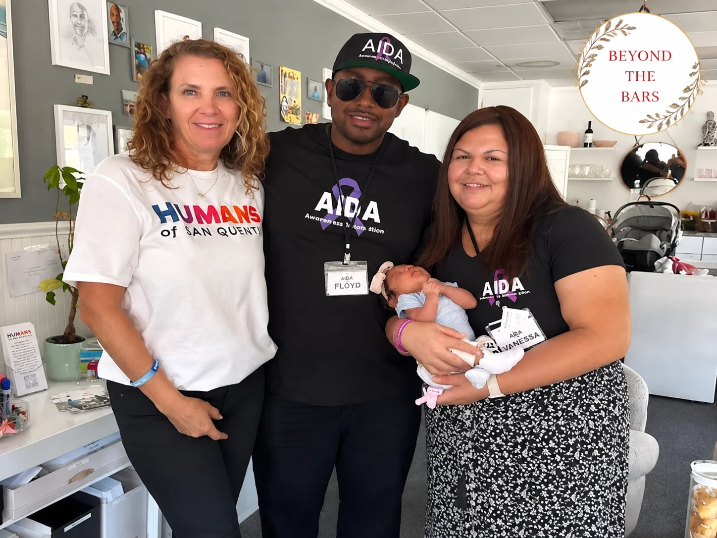 Three people, including a man named Floyd, stand together in an office. Floyd wears a black AIDA t-shirt and a matching cap, holding a baby. The woman on the left wears a white 'Humans of Ian Quint' shirt.