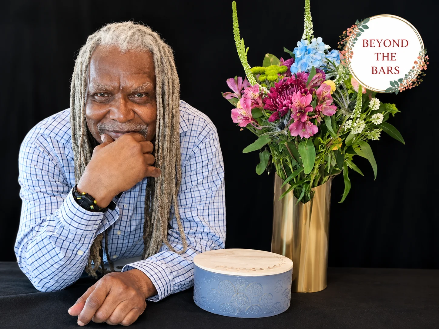 Gerald, a man with long grey dreadlocks, rests his chin on his hand while looking at a floral arrangement in a gold vase and a decorative box.