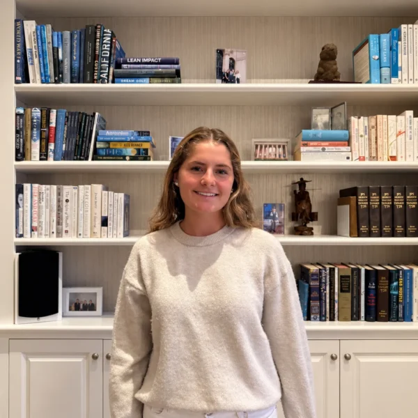 Our Team 51 Hyla with shoulder-length, wavy light-brown hair stands in front of a built-in white bookshelf filled with books, framed photographs, and decorative sculptures. She wears a cream knit sweater and light-colored trousers in a warmly lit, cozy library setting.