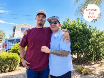 Two men stand side-by-side outdoors, smiling at the camera. The man on the left wears a maroon t-shirt and a tan cap, while the man on the right wears a light blue shirt, sunglasses, and a blue cap. They have their arms around each other's shoulders.