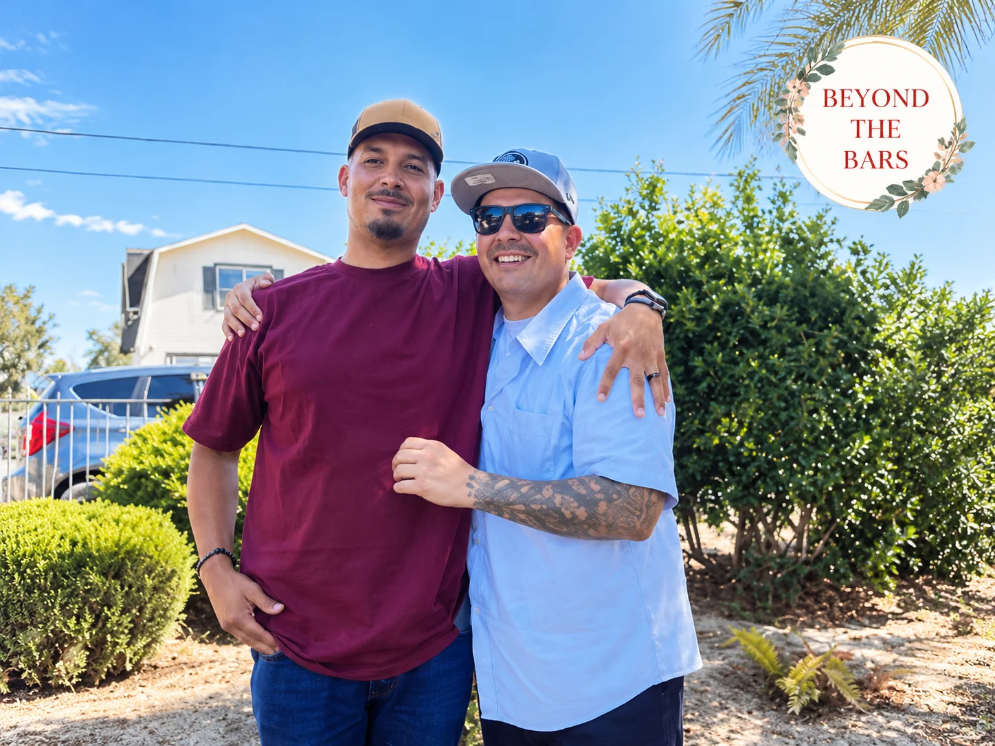Two men stand side-by-side outdoors, smiling at the camera. The man on the left wears a maroon t-shirt and a tan cap, while the man on the right wears a light blue shirt, sunglasses, and a blue cap. They have their arms around each other's shoulders.
