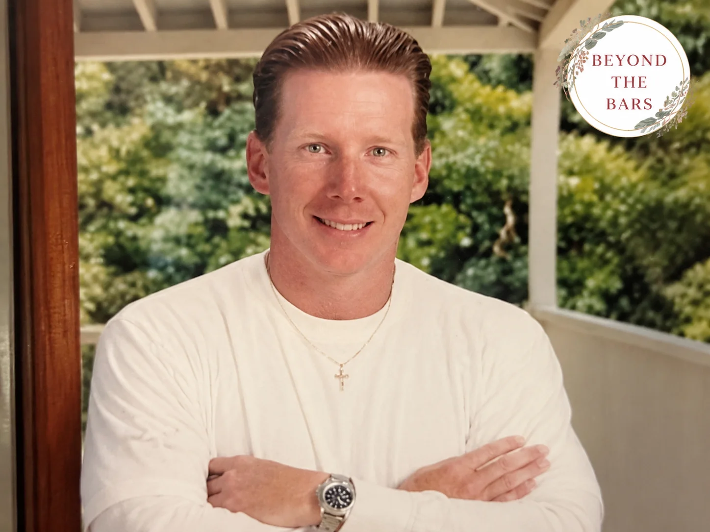 Mark, a man with short hair, wearing a white shirt and a cross necklace, stands with his arms crossed in front of a window.