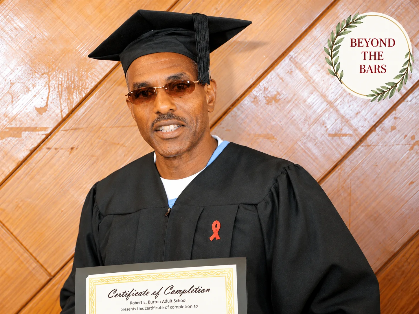 A man in a black graduation cap and gown with a red awareness ribbon, holding a certificate from Robert E. Burton Adult School.