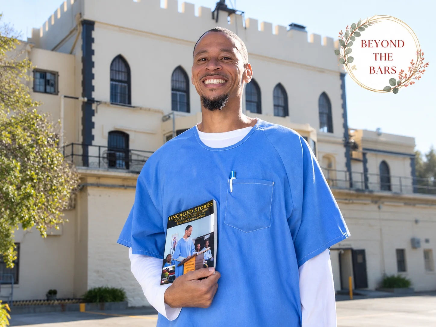 A smiling man in blue scrubs holds a book titled 'Uncaged Stories' in front of a historic white building.