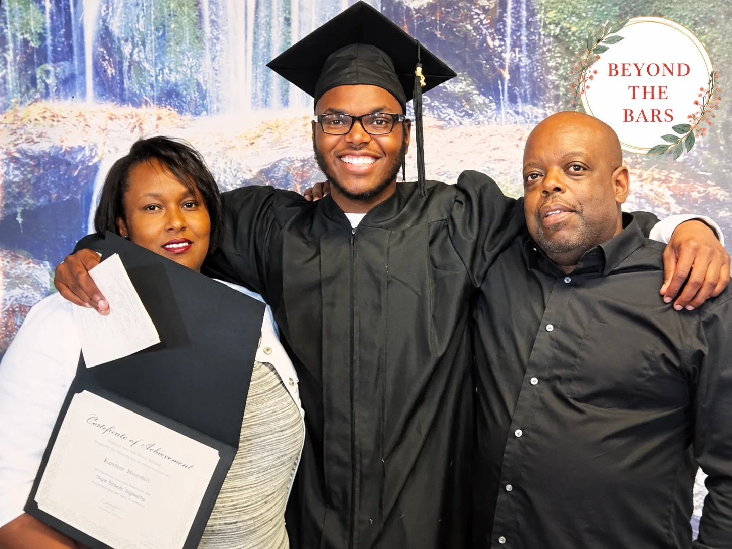A graduate in a black cap and gown stands with his arms around two smiling adults, one holding a certificate of achievement, in front of a waterfall backdrop.