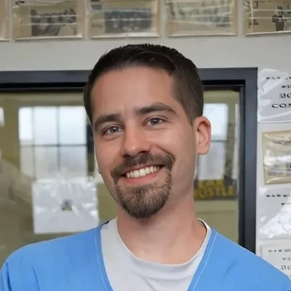Our Team 23 A smiling man with a beard and blue scrubs stands in a workshop with technical drawings on the wall.