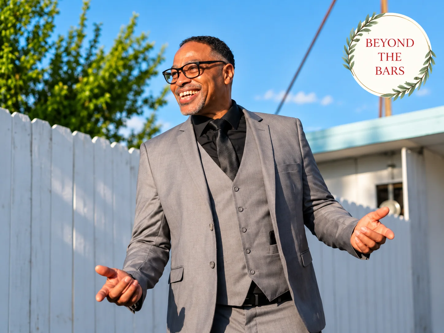 A man in a grey suit and glasses smiles with open arms outdoors.