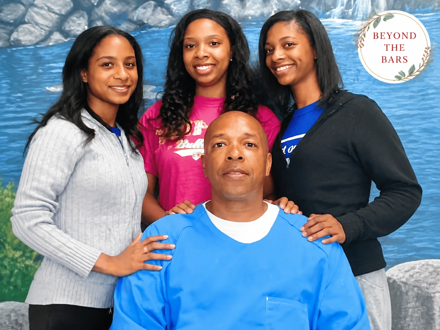 A man in a blue shirt sits in front of three smiling women who have their hands on his shoulders, with a blue waterfall mural in the background.