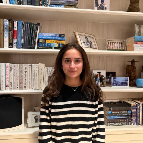 Our Team 70 Young woman with long, wavy brown hair wearing a black-and-white striped sweater, standing in front of a built-in bookshelf filled with books, framed family photographs, and decorative keepsakes.