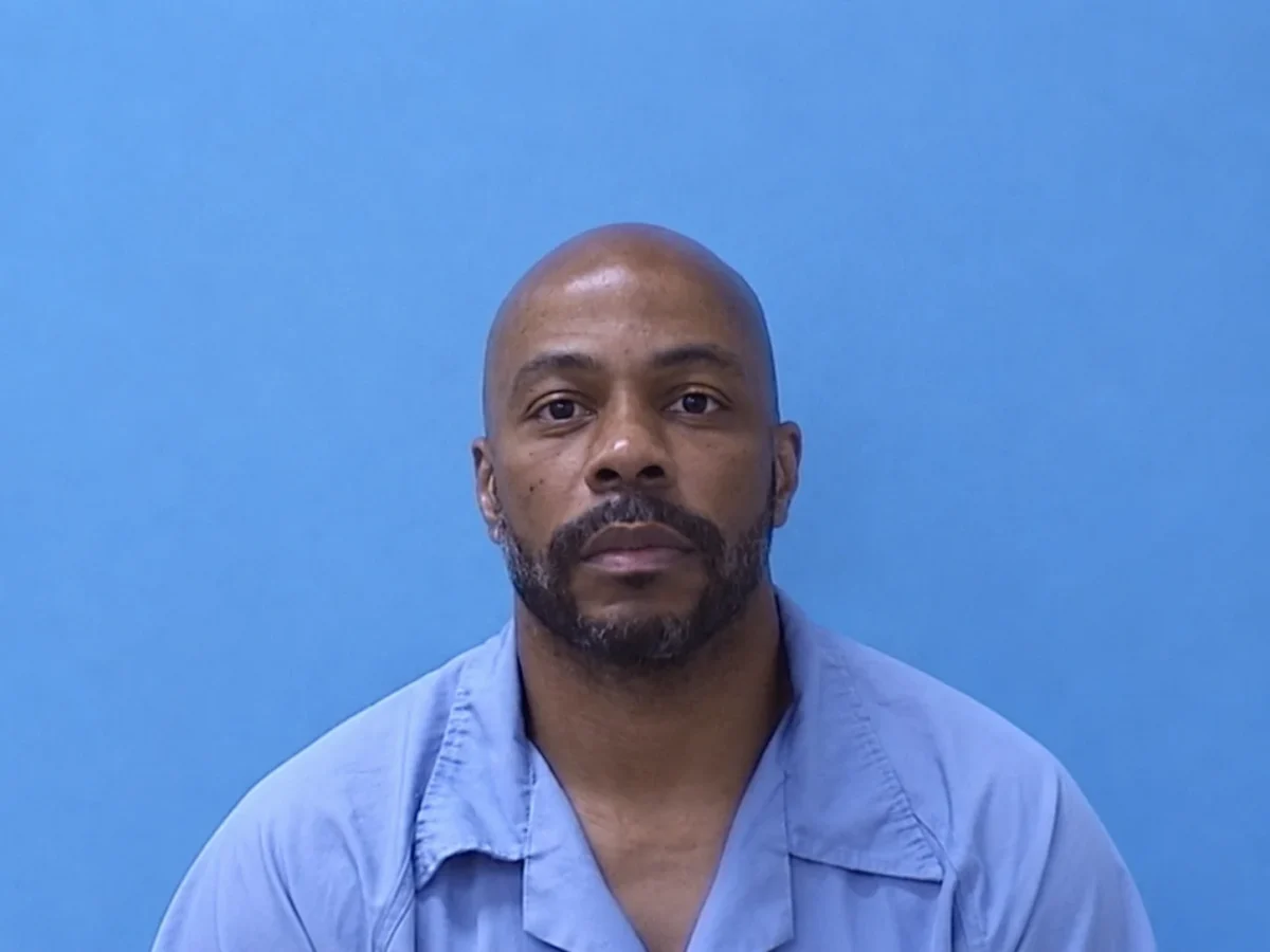 Portrait of a Chicago man with a shaved head and neatly trimmed beard, wearing a light blue shirt against a blue background, looking directly at the camera with a calm, serious expression.