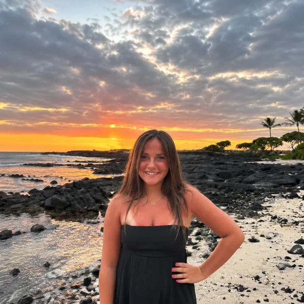 Our Team 69 A smiling young woman with long brown hair wearing a black strapless dress poses with one hand on her hip on a rocky beach at sunset. The ocean, dark volcanic rocks, palm trees, and a dramatic cloudy sky glowing orange and gold create a scenic tropical backdrop.