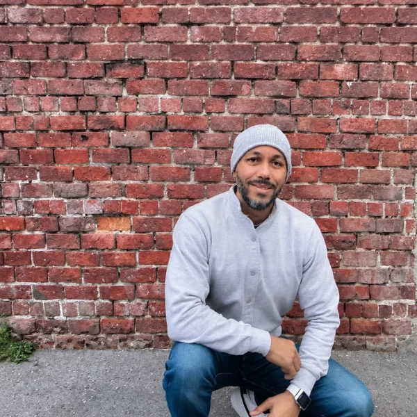 Our Team 25 Outdoor portrait of a man crouching beside a red brick wall, wearing a gray beanie, light shirt, blue jeans, and white sneakers.
