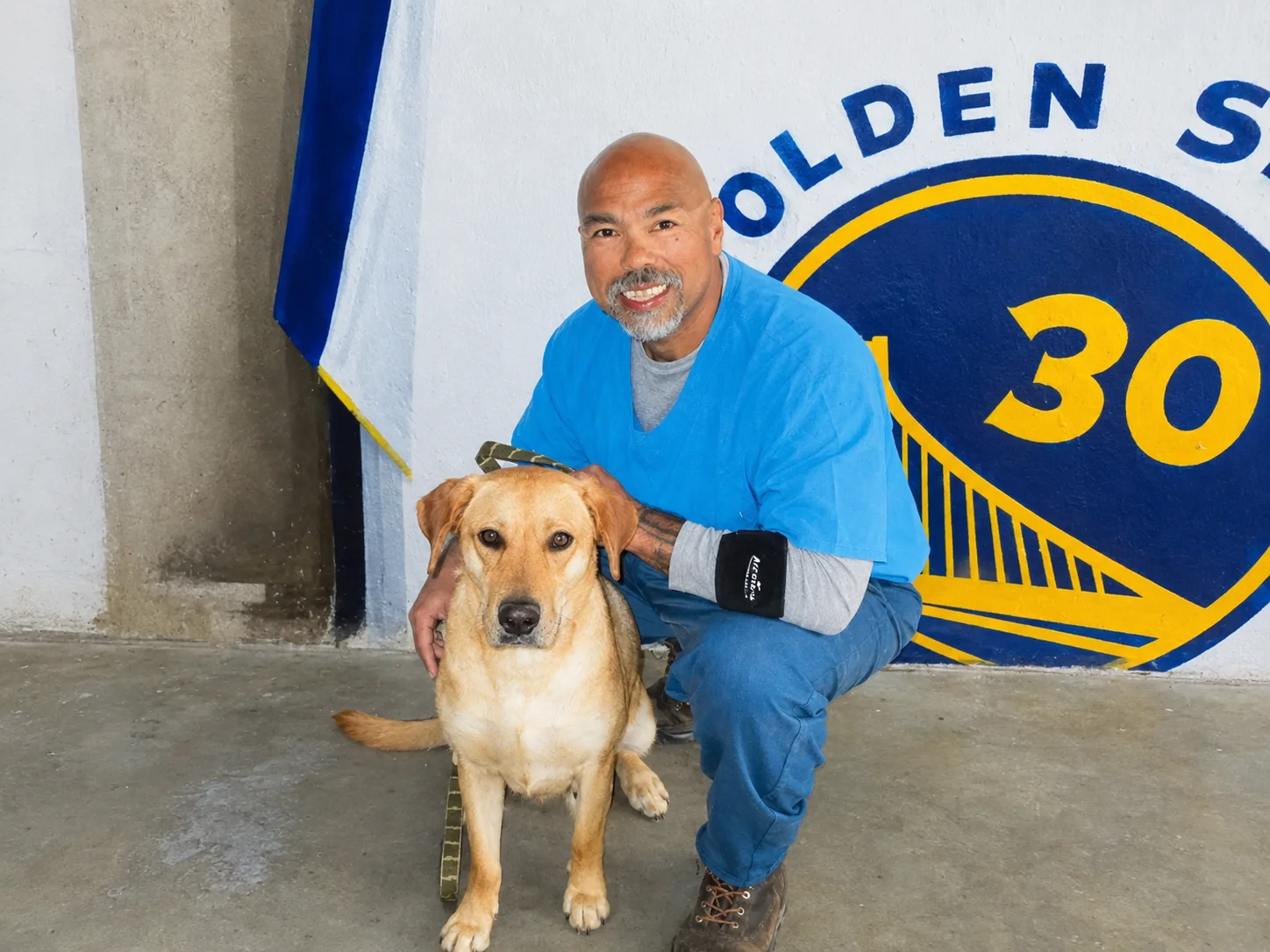 Joseph, a smiling incarcerated man in blue clothing crouches beside a tan dog in a prison dog program, highlighting rehabilitation and personal growth.
